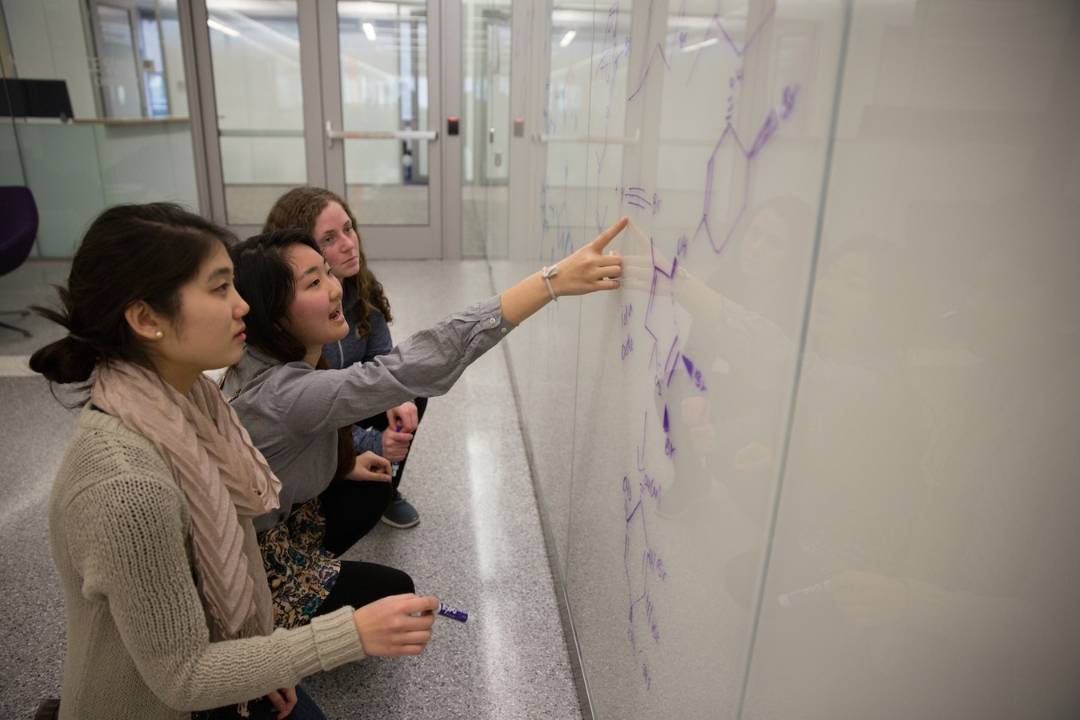 Three women standing in front of a whiteboard with chemistry work on it