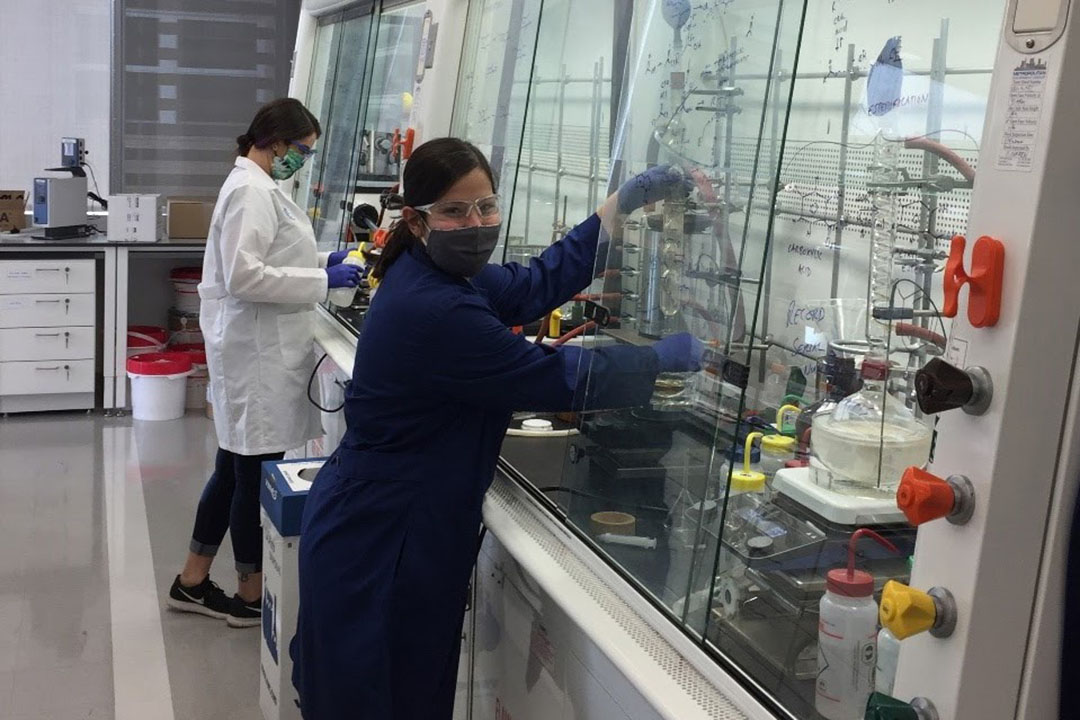 A woman dressed in lab coat and goggles works in a lab surrounded by equipment